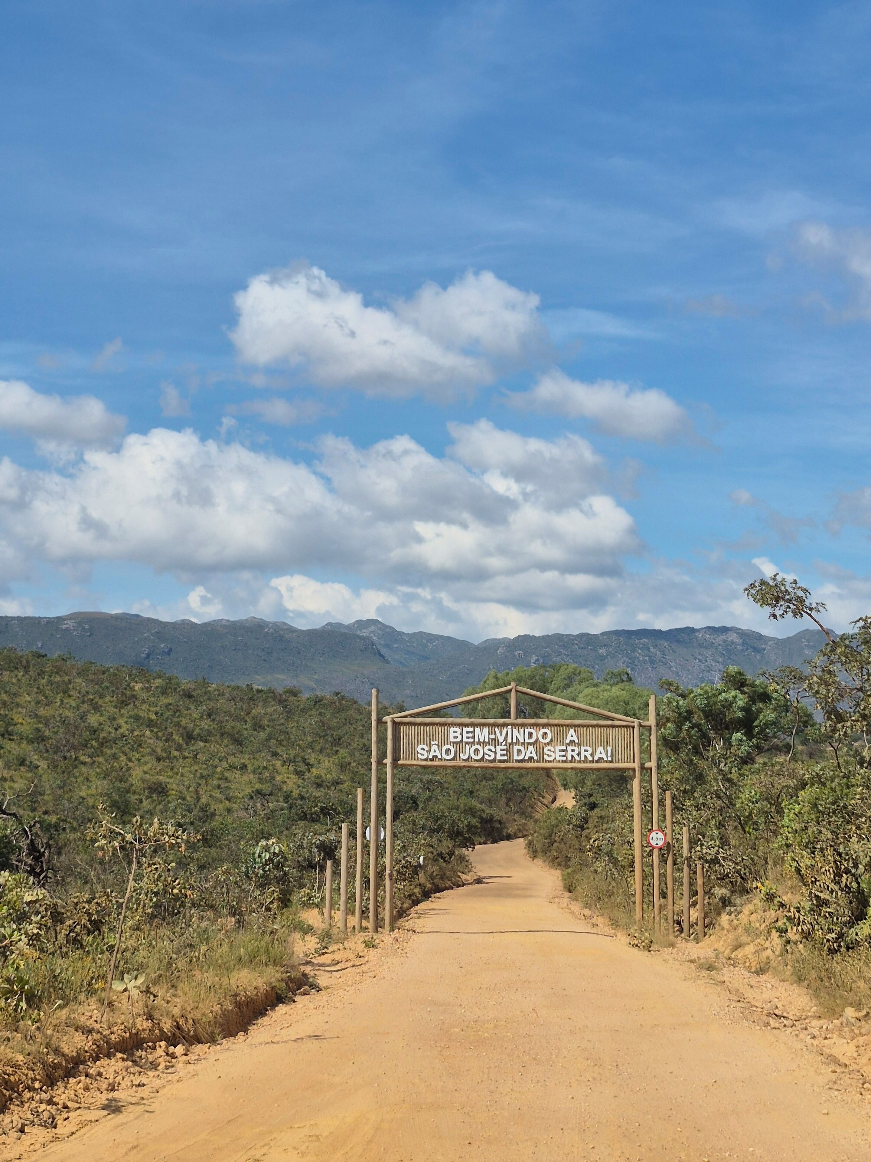 Uma estrada de terra batida que leva a uma placa com a inscrição 'Bem-vindo a São José da Serra!'.