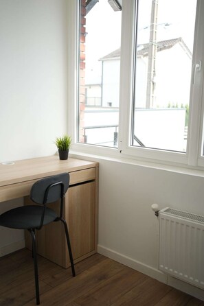 Et værelse med et træskrivebord, en sort stol, en potteplante, et vindue og en radiator.