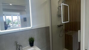 Et moderne badeværelse med glasvæg ved brusebad, en hvid vask og et spejl med indbygget lys.