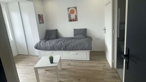 Et soveværelse med en seng, et lille bord med en plante, et billede på væggen og en dør.
