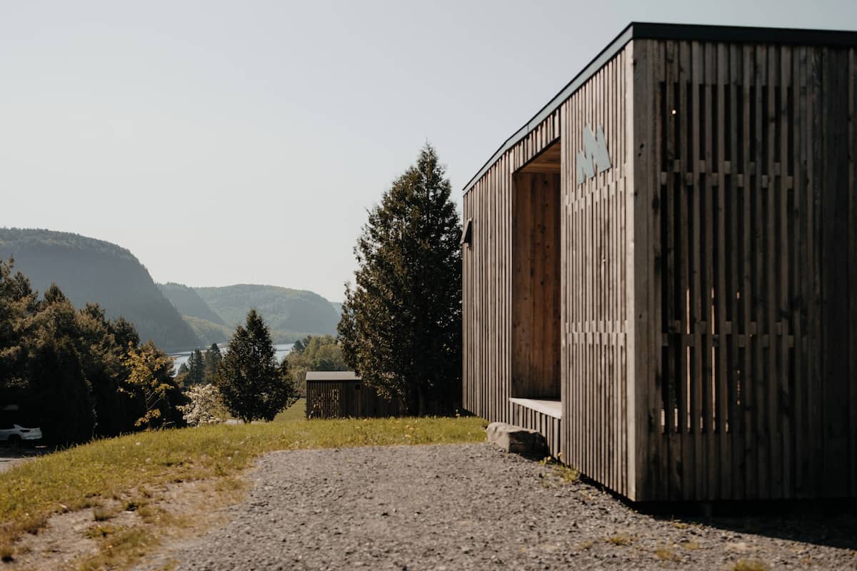A wooden building with a sloped roof, a gravel path, and a view of a lake and mountains in the background.