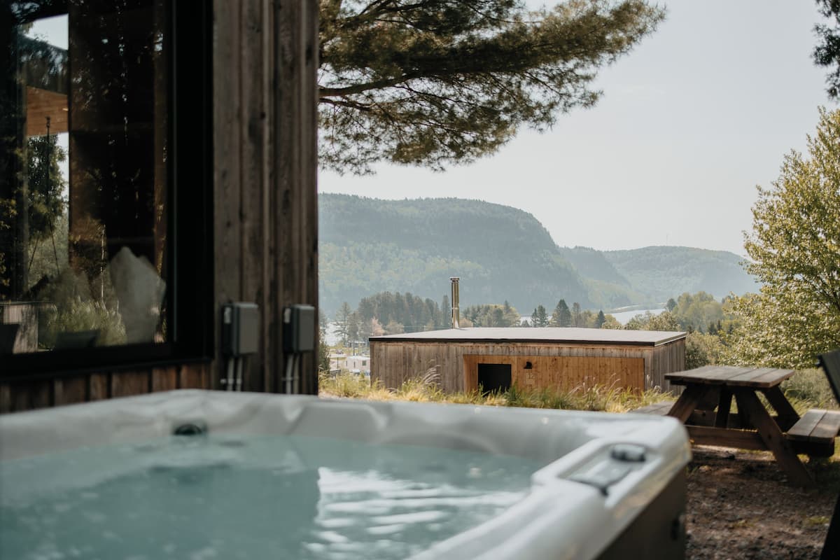 A hot tub with a wooden deck, a wooden building, and a view of a lake and mountains.