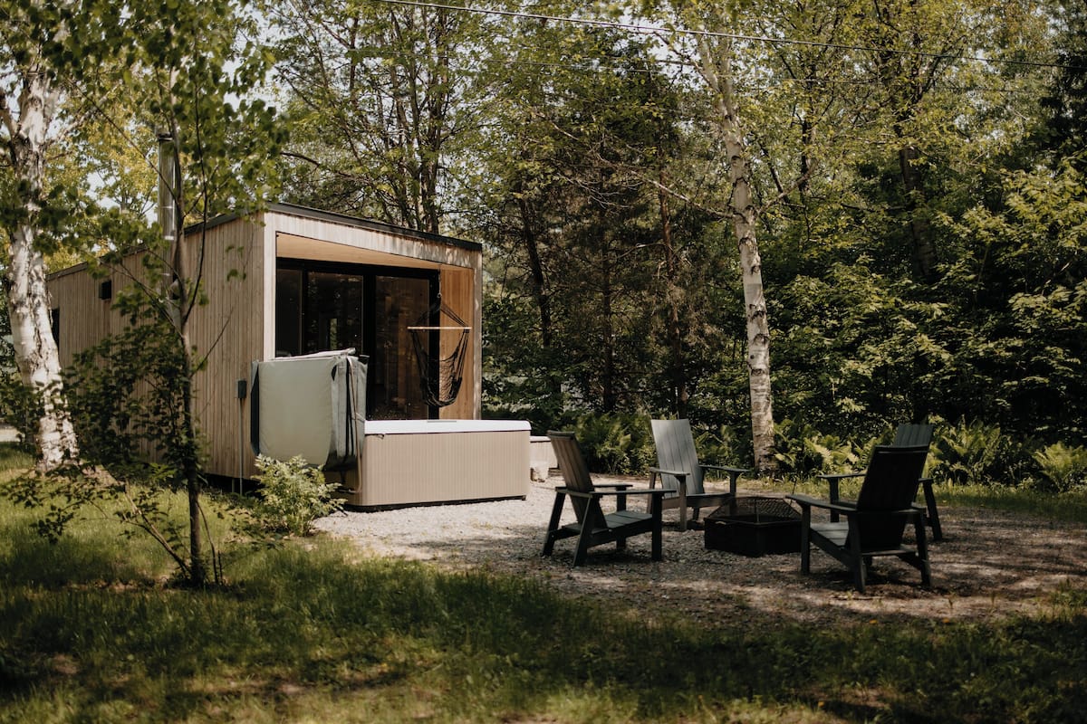 A small wooden cabin with a glass door, surrounded by trees, and furnished with outdoor seating.