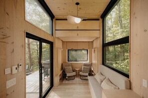 A cabin interior with a wooden floor, a sofa, two chairs, a small table, and large windows offering a forest view.