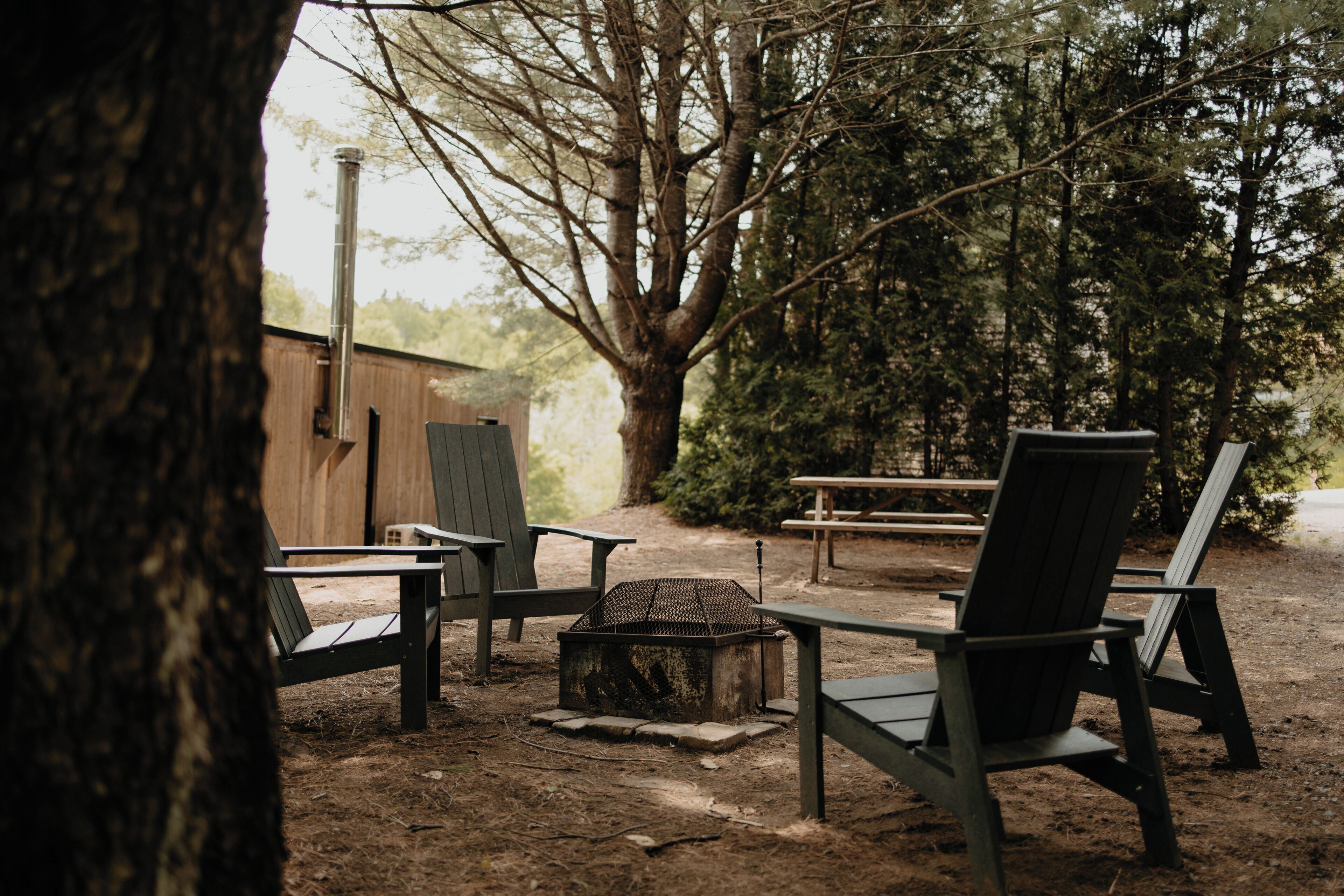 A rustic outdoor seating area with Adirondack chairs, a fire pit, and a wooden structure in the background.