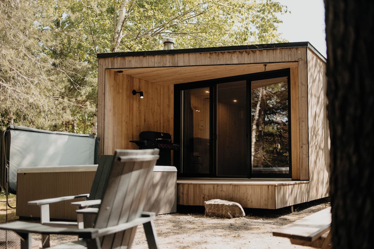 A wooden cabin with a large window, a small outdoor seating area, and a grill.