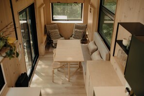 Interior de autocaravana compacto y moderno, con revestimientos de madera, una mesa de comedor, dos sillones acolchados, un sofá y ventanas amplias que dejan entrar luz natural.