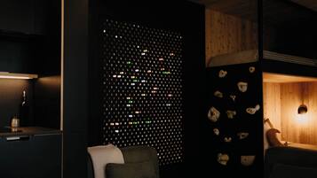 A room with a dark wall featuring a grid of small, colorful lights, a wooden ceiling, a bed with a headboard, and a small table with a lamp.