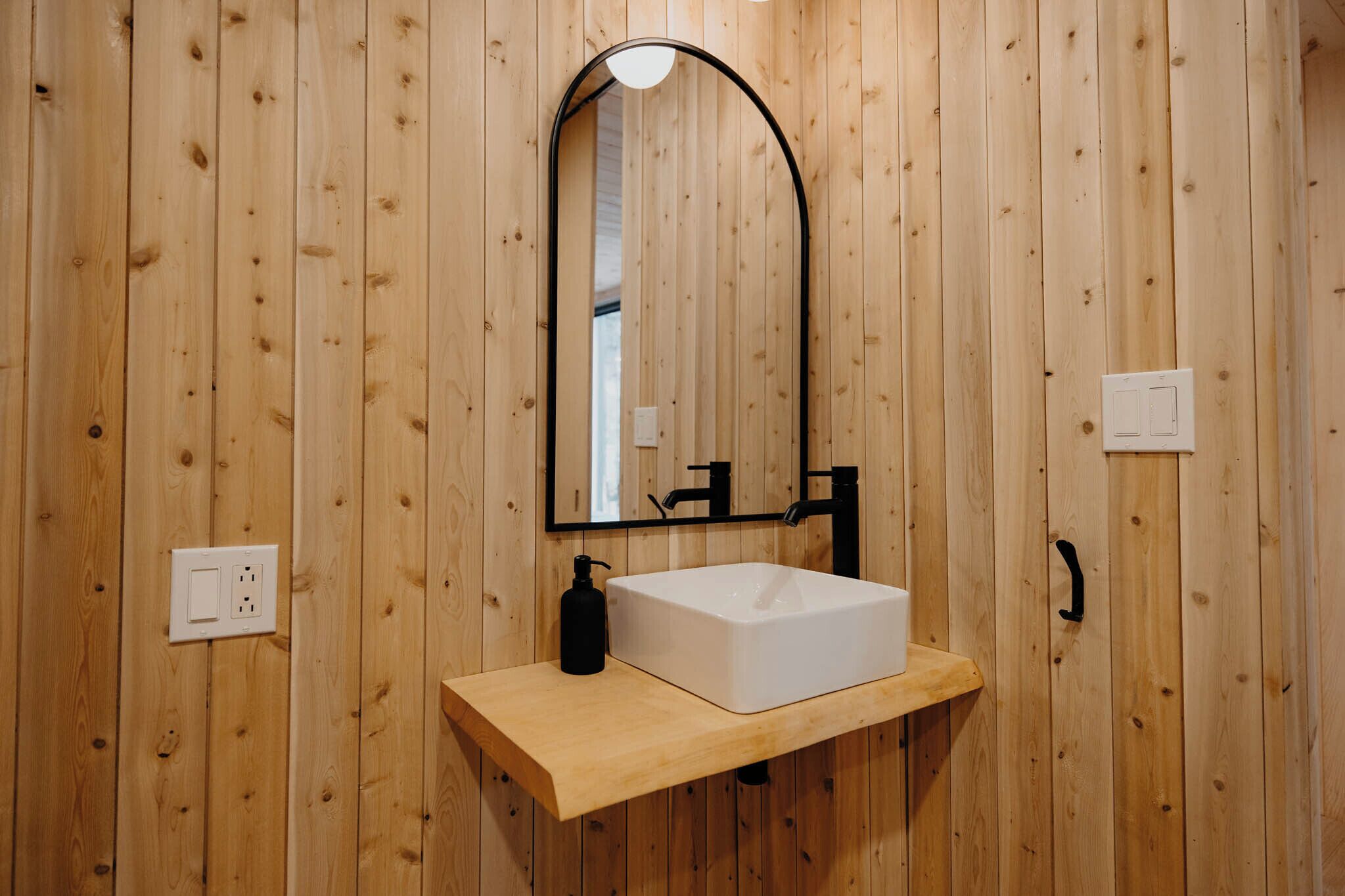 Une salle de bain aux murs lambrissés, avec un lavabo blanc, un robinet noir et un miroir dont le sommet est incurvé.
