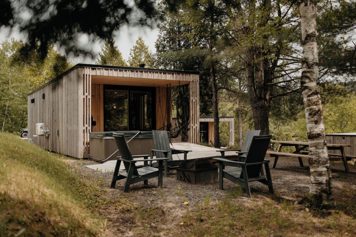 A wooden cabin with a deck, surrounded by birch trees, featuring outdoor seating and a picnic table.