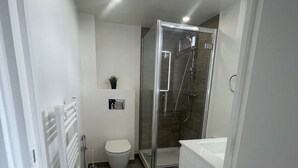 Un baño estrecho con inodoro, ducha con mampara de vidrio y bañera blanca.
