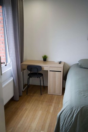 Ein Schlafzimmer mit einem Holztisch, einem Stuhl, einer kleinen Pflanze und einem Bett mit grauer Decke.