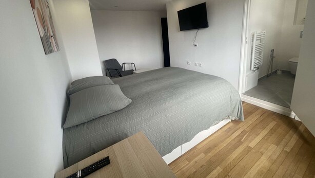 Un dormitorio con una cama, una silla, un televisor montado en la pared y un baño a la vista a través de una puerta abierta.