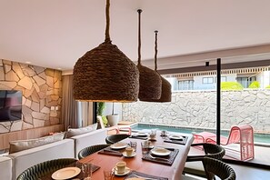 A modern dining area with a stone wall, a pool, and a view of a building. - Cond Triplex - By MME (Jacuípe)
