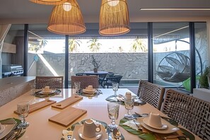 A dining area with a table set for four, large windows, and a stone wall in the background. - Cond Triplex - By MME (Jacuípe)