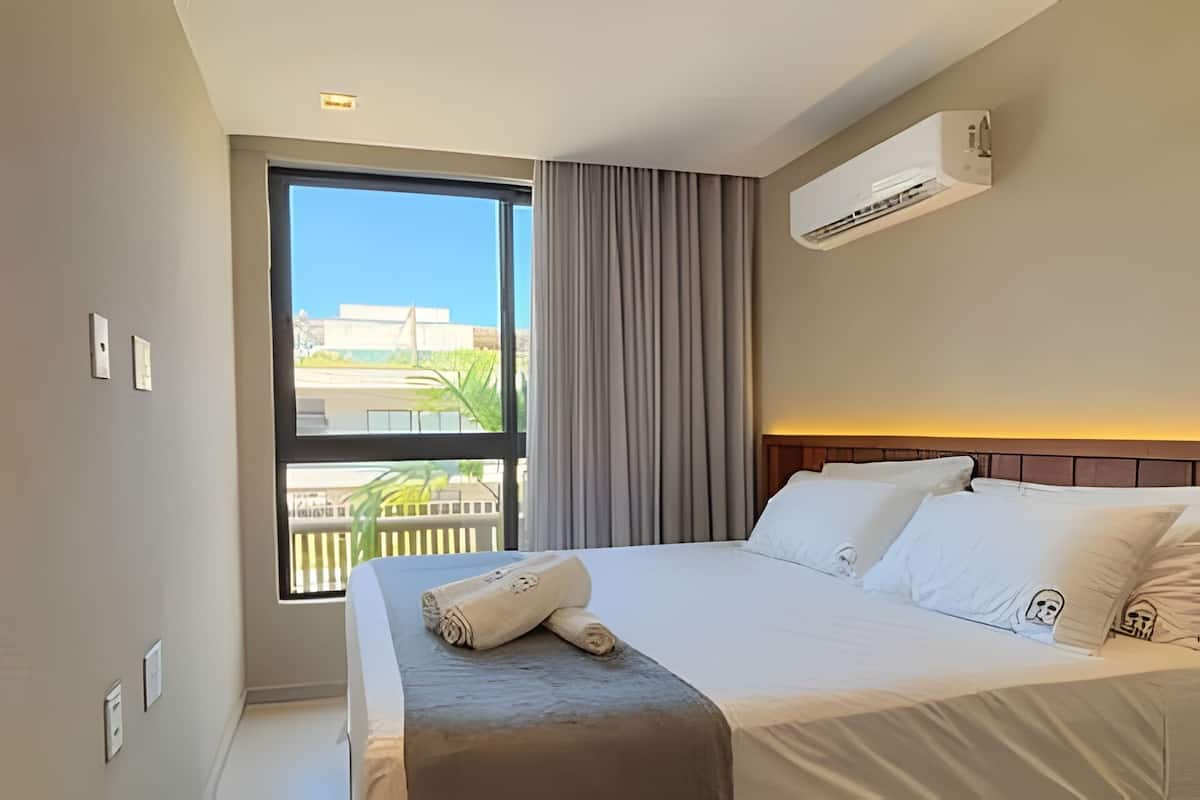 A hotel room with a bed, a window with curtains, an air conditioning unit, and a view of buildings outside.