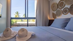 A bed with a textured cover, two hats, a window with a view of palm trees, and a wall with woven wall art.