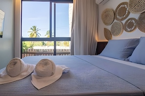 A bed with a textured cover, two hats, a window with a view of palm trees, and a wall with woven wall art.