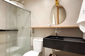 A modern bathroom with a glass shower enclosure, a white toilet, a black sink, and a round mirror. - Cond Triplex - By MME (Jacuípe)