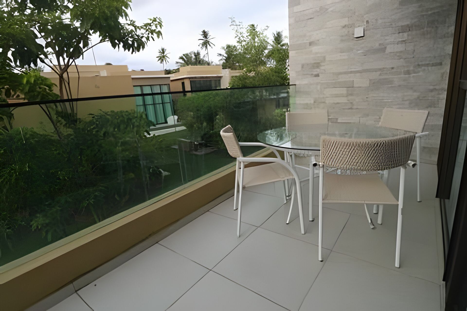 A balcony with a glass table and chairs, a view of greenery and buildings, and a tiled floor.