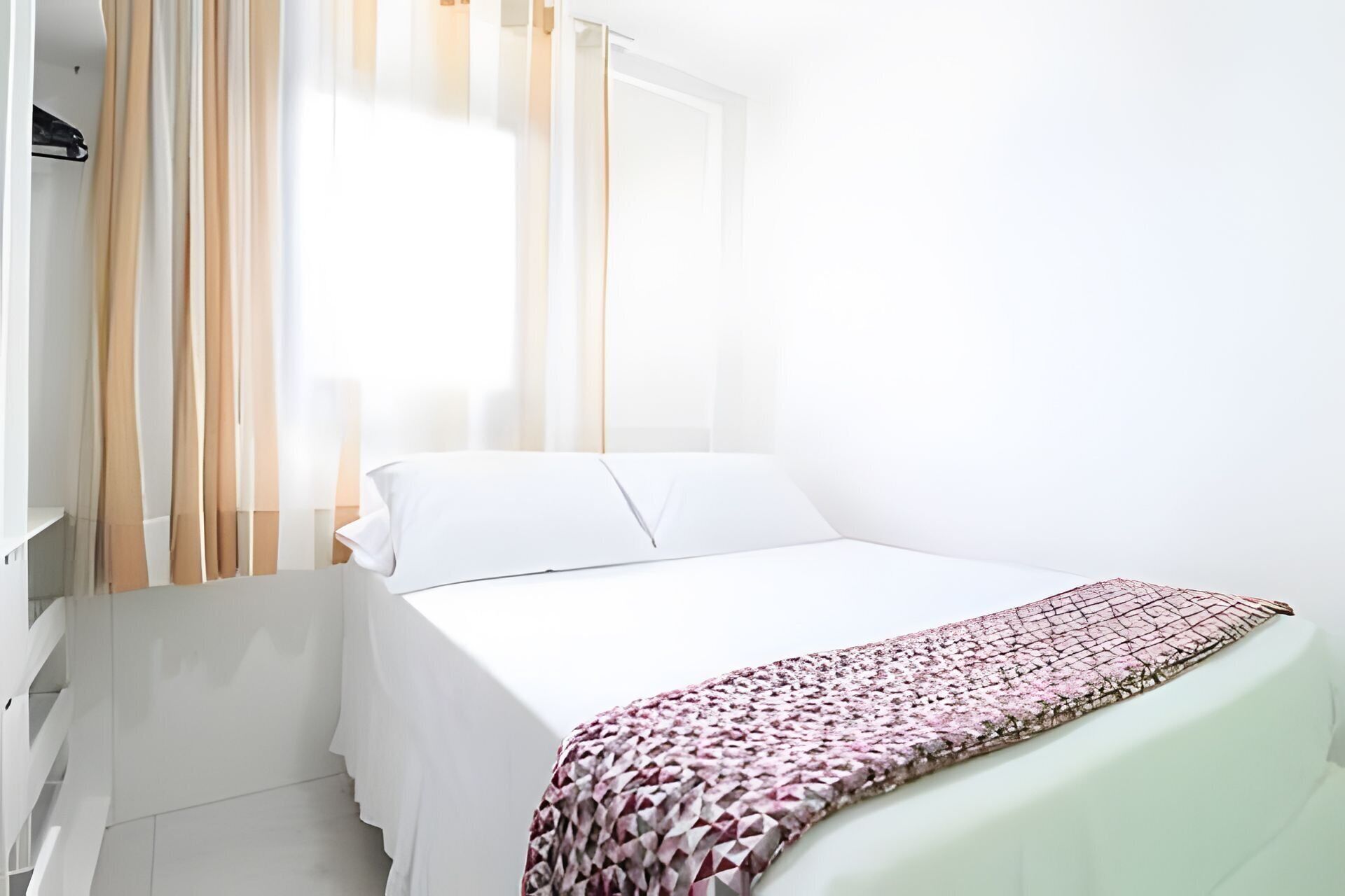 A single bed with white bedding, a patterned throw, and sheer curtains.