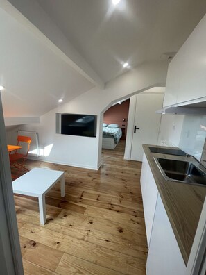 Eine moderne Küche mit Backofen, ein Essbereich mit weißem Tisch und orangefarbenen Stühlen sowie ein Schlafzimmer mit Bett und Fernseher.