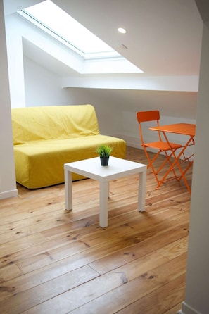 Een kamer met een gele bank, een witte tafeltje met een plant, oranje stoelen en een dakvenster.