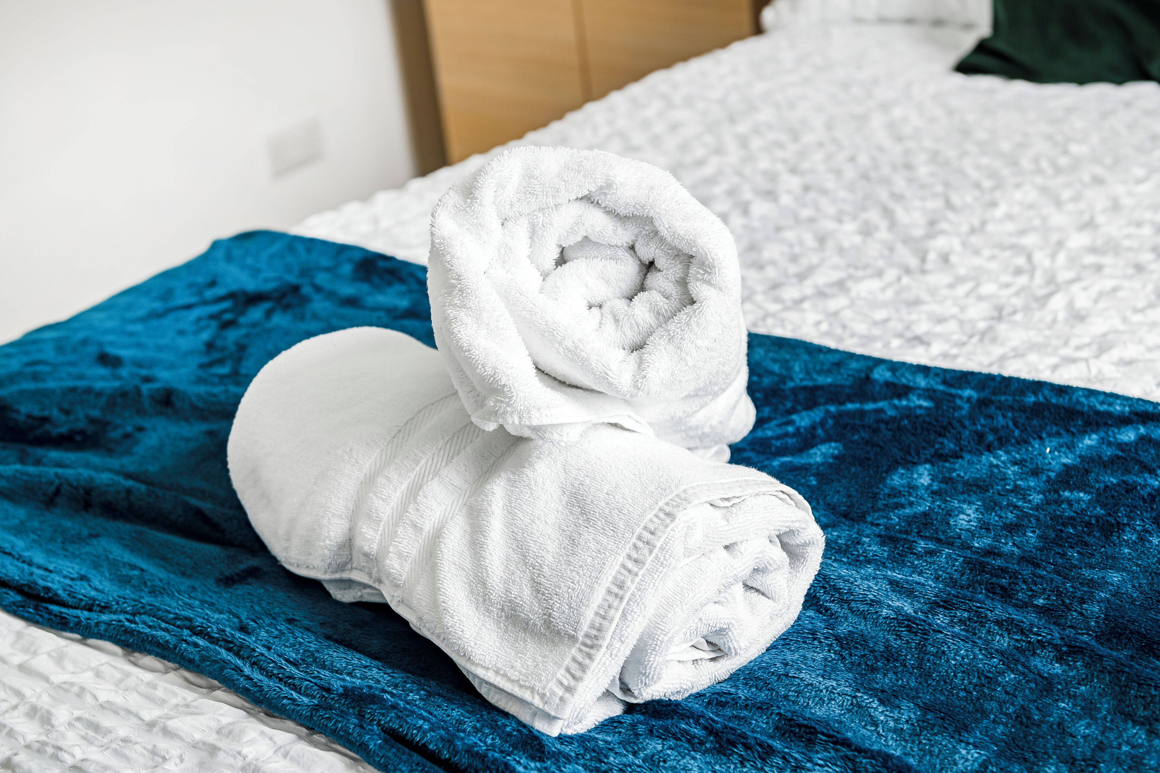 Ein ordentlich gefaltetes weißes Handtuch auf einer blauen, strukturierten Decke auf einem Bett mit einer weißen, gesteppten Bettdecke.