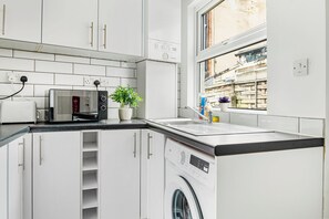 A compact kitchen with a washing machine, a microwave, and a window with a view of a building. - NEW! 5BR House, free parking, near City Centre (Nottingham)