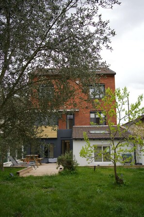 A modern two-story building with a flat roof, a small garden area with a wooden table and chairs, and a tree in the foreground. - Cozy Private Room | Coliving 30min from Paris St-Lazare (Argenteuil)