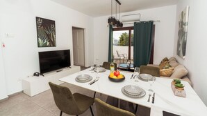 A dining area with a TV, artwork, and a table set for four with plates, glasses, and a fruit bowl. - SerenityOne - Centrally located Apt,SO19 (Agia Napa)