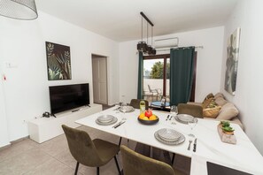 A dining area with a TV, artwork, and a table set for four with plates, glasses, and a fruit bowl. - SerenityOne - Centrally located Apt,SO19 (Agia Napa)