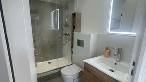 Moderni kylpyhuone, jossa on lasiseinällä varustettu suihku, wc ja peilillä varustettu pesuallas.