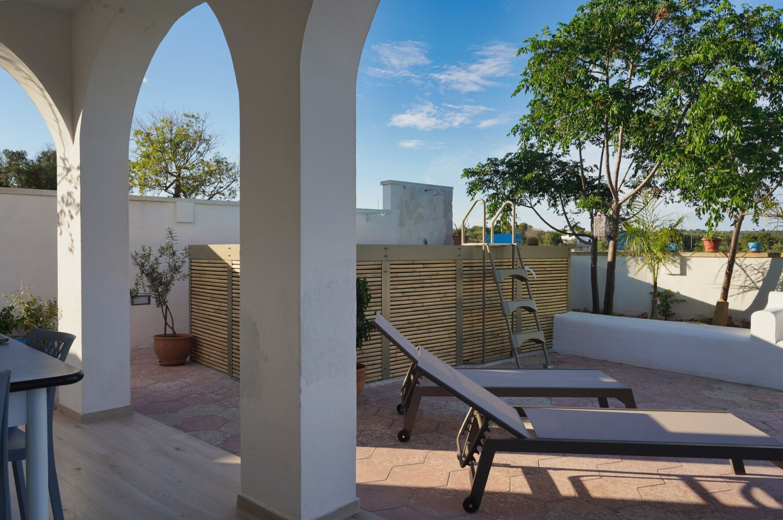 Eine Terrasse mit Tisch und Stühlen, ein Liegestuhl, Kübelpflanzen und ein Holzzaun.
