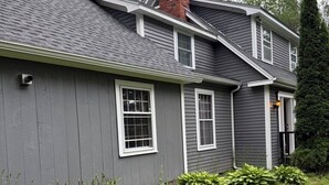 A two-story house with a grey exterior, a brick chimney, and a small front yard with green plants. - Spacious Family Getaway Near Sunapee & Dartmouth (Croydon)
