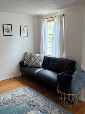 Ein Wohnzimmer mit einem schwarzen Ledersofa, einem gemusterten Zierkissen, einem blau-weißen Teppich, einem Korbwannen und zwei gerahmten floralen Kunstwerken an der Wand.