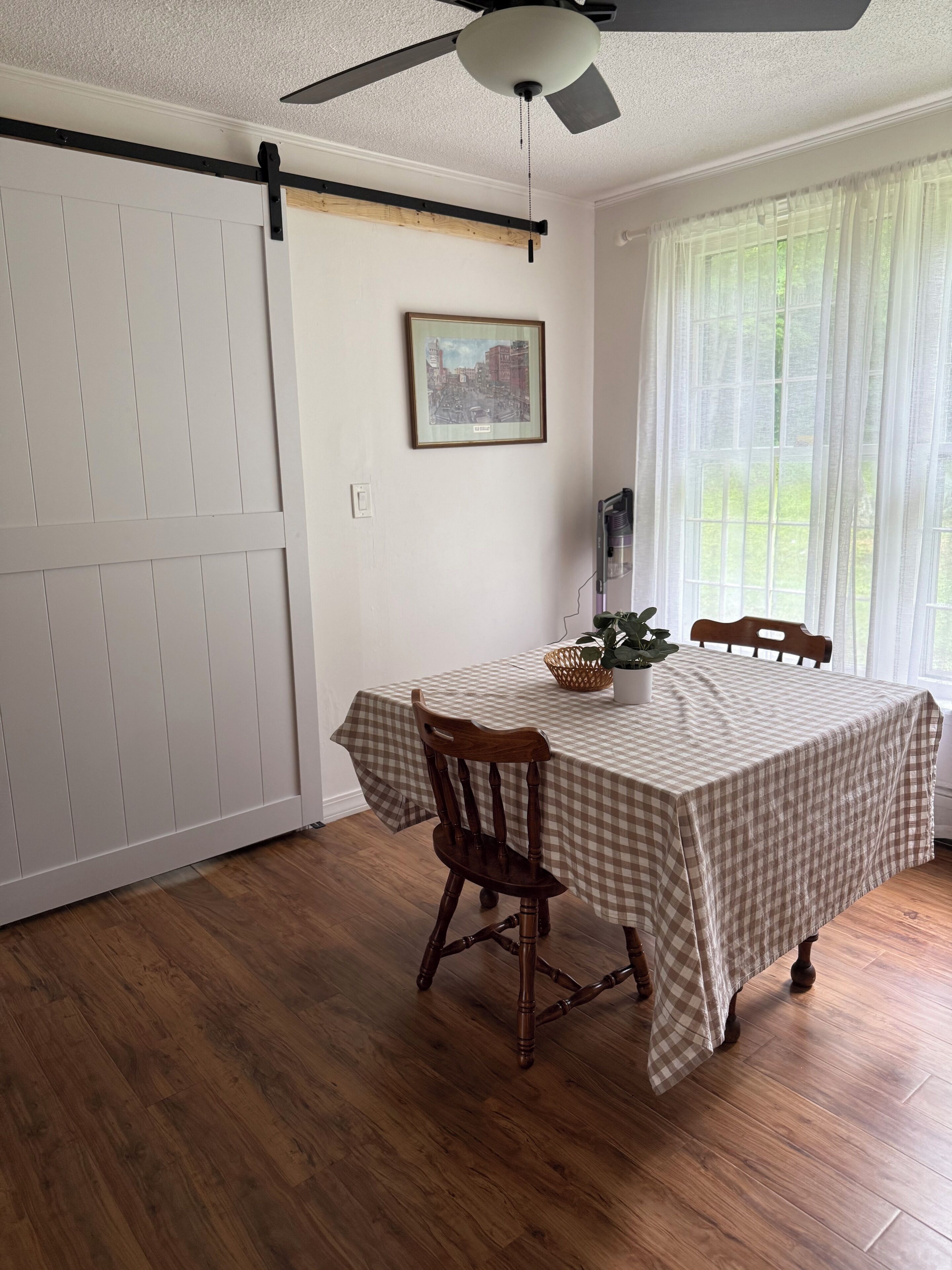 A dining area with a checkered tablecloth, wooden chairs, a ceiling fan, a framed cityscape picture, and a window with sheer curtains.