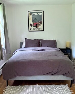 Ein Schlafzimmer mit Bett, Nachttisch, Lampe, gerahmtem Poster an der Wand und einem Fenster mit Vorhängen.