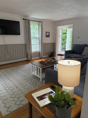 Ein Wohnzimmer mit einer Couch, einem Couchtisch mit Buch und Pflanze, einem an der Wand montierten Fernseher und einem Fenster mit Vorhängen.