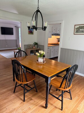 Ein Esszimmer mit Holztisch, schwarzen Stühlen, einer Chandelierleuchtre und einer dahinterliegenden Küche.