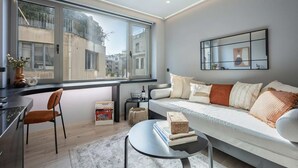 Una sala moderna con un sofá, una mesa de centro, una silla, una mesita auxiliar y un amplio ventanal con vistas a edificios.
