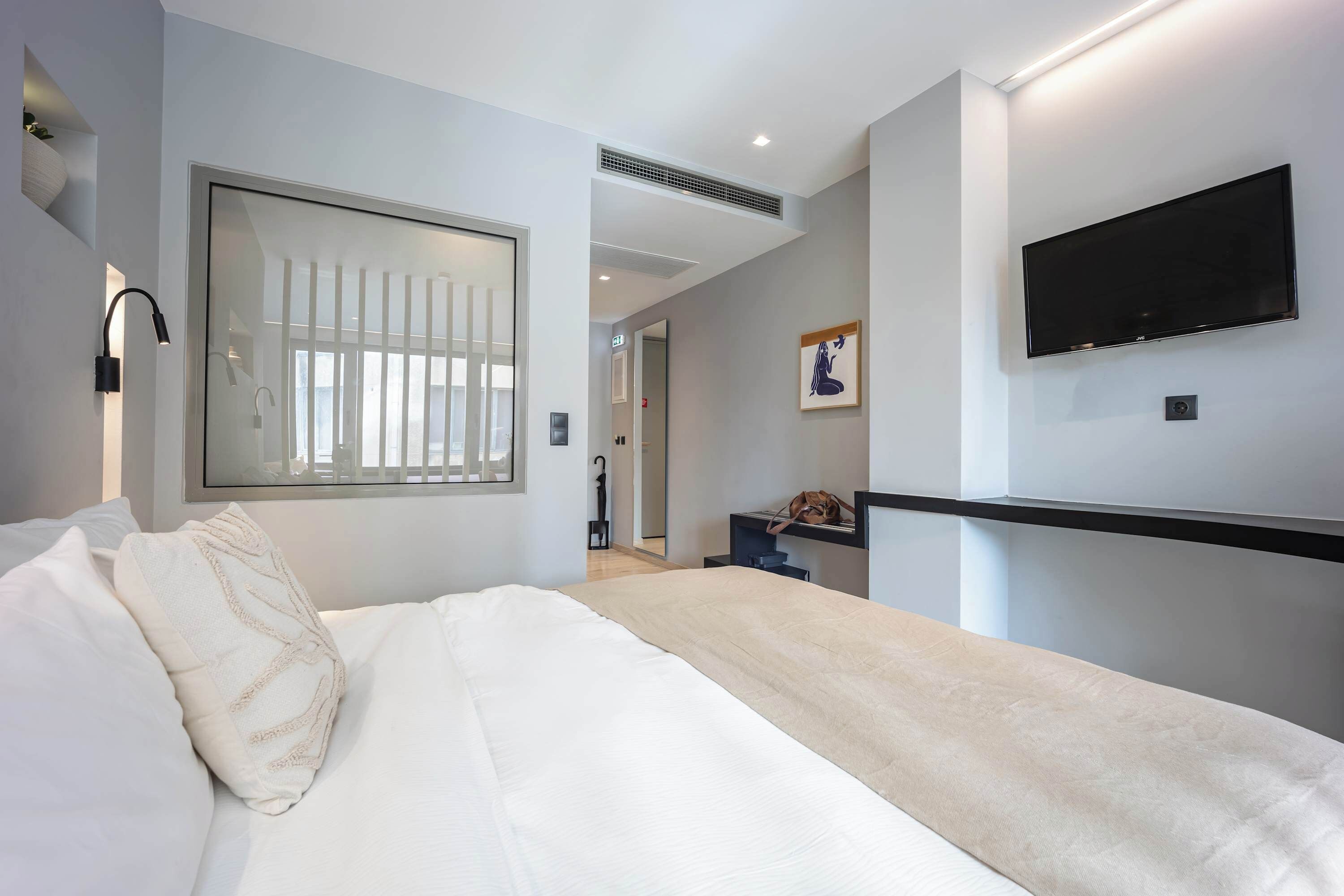Une chambre d’hôtel moderne dotée d’un grand lit, d’un téléviseur à écran plat fixé au mur, d’une table de chevet et d’une fenêtre avec des stores.