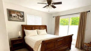 Une chambre à coucher avec un lit en bois, une table de chevet, un ventilateur de plafond, une fenêtre avec des rideaux et une chaise.