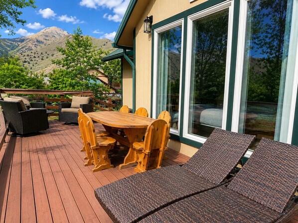 Um terraço com mesa e cadeiras de madeira, um sofá e uma janela grande com vista para as montanhas.