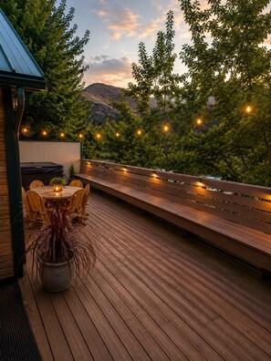 Um deck externo com mesa e cadeiras de madeira, luzes de festa e vista para uma paisagem montanhosa.