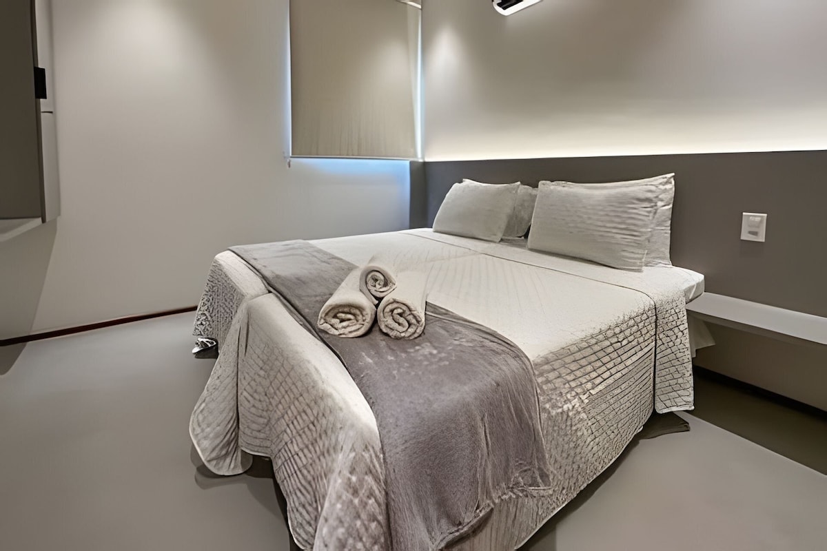 Quarto moderno com uma cama grande, mesa de cabeceira, espelho e luminária na parede.