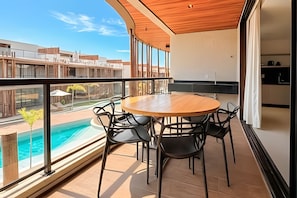 A balcony with a wooden table and chairs, a pool view, and a cityscape in the background.