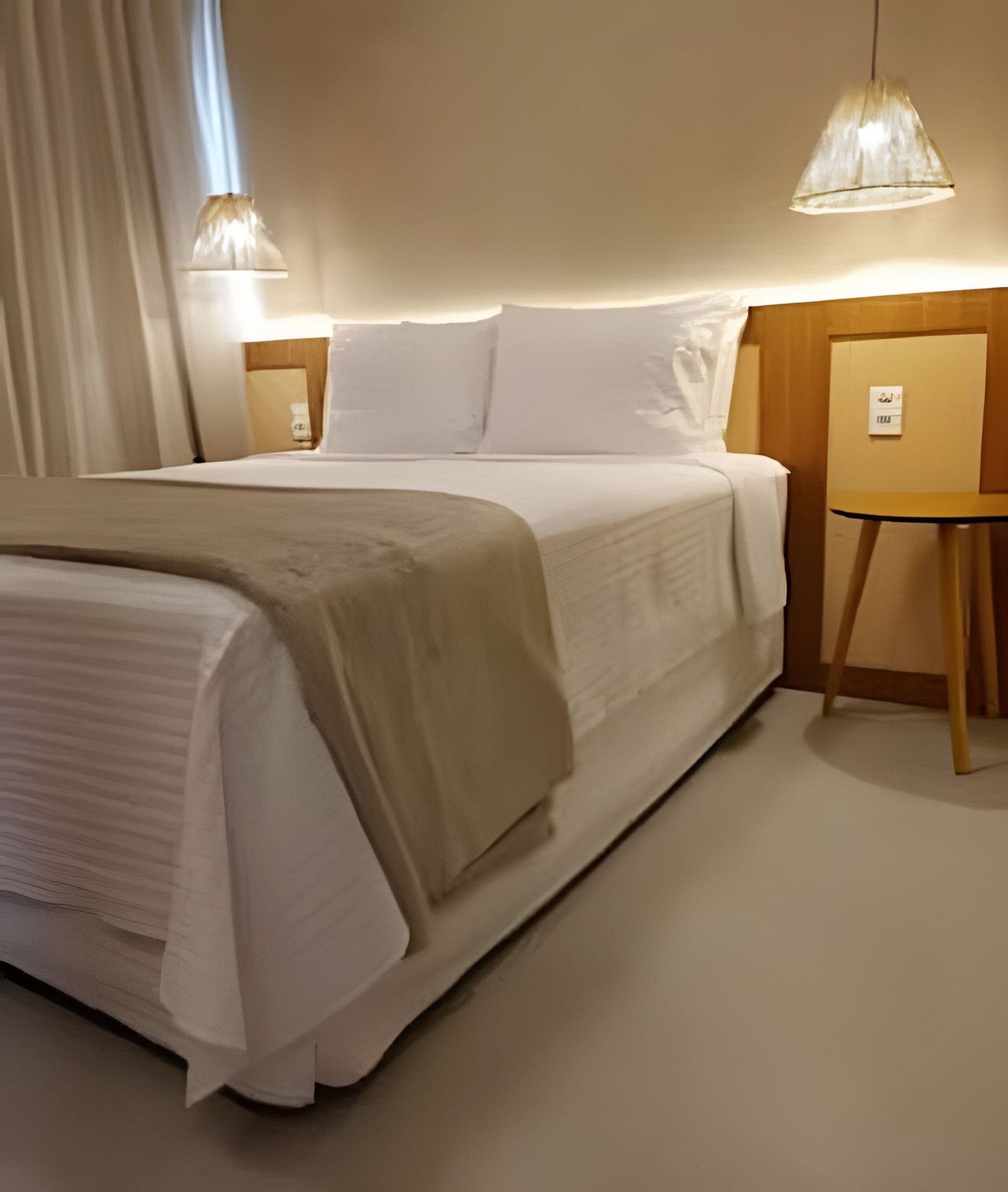 A neatly made bed with a white comforter and a beige throw blanket, two bedside lamps, and a small wooden nightstand with a sign.
