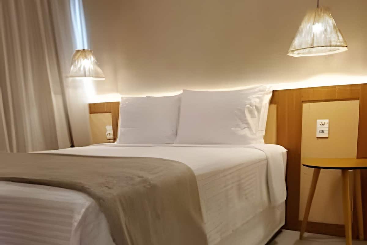 Un lit bien fait, avec une couette blanche et un plaid beige, deux lampes de chevet et une petite table de nuit en bois sur laquelle est posé un petit panneau.
