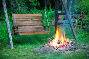 Eine hölzerne Bank schwebt über einem Lagerfeuer in einer Wiese.